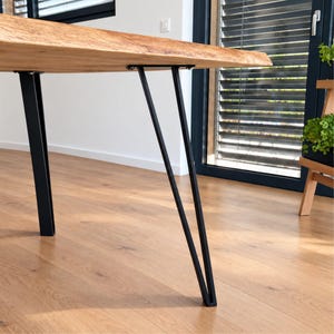 May include: A live edge wooden table top featuring a natural light brown grain pattern, supported by two black metal V-shaped legs attached to the underside with visible screws. The table surface has a smooth, unfinished appearance.