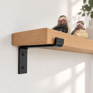 Op de afbeelding: Een plank van lichtgekleurd natuurhout gemonteerd aan een muur met een matzwarte metalen L-vormige beugel. De plank heeft een zichtbare houtnerfstructuur en bevat twee kleine, decoratieve glazen vogelbeeldjes met gespikkelde patronen.