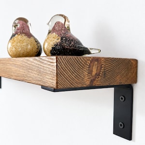 Op de afbeelding: Een rechthoekige houten plank met zichtbare houtnerf en een noest, ondersteund door twee zwarte metalen L-vormige beugels vastgezet met schroeven. Twee glazen vogelbeeldjes met gespikkelde roze, gouden en zwarte patronen staan op de plank.
