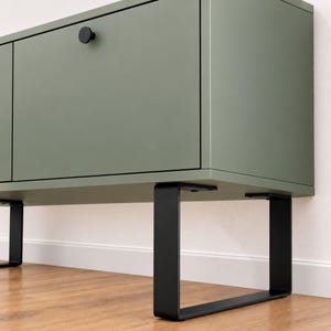 Op de afbeelding: Een saliegroene dressoirkast met twee deuren en kleine, ronde, matzwarte knoppen. De kast wordt ondersteund door twee U-vormige, zwarte metalen poten die een stevige basis vormen voor de rechthoekige opbergkast.