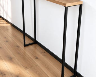 Peut inclure: Une console haute et étroite dotée d'un plateau rectangulaire en bois brun clair avec un grain visible. La table est soutenue par une fine structure en métal noir composée de deux pieds rectangulaires reliés par une barre horizontale.