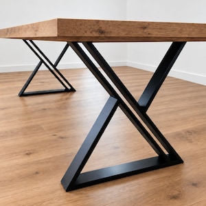 Puede incluir: Una mesa rectangular con un tablero grueso de madera marrón claro con vetas, nudos y textura visibles. La mesa se apoya sobre dos patas de metal negro, cada una construida con un marco geométrico en forma de X sobre una base plana.