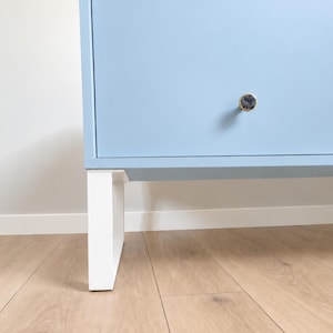 Op de afbeelding: Een lichtblauwe kast met een enkele lade en een ronde, goudkleurige knop. De kast wordt ondersteund door een witte, U-vormige metalen poot. Het oppervlak van de kast is glad en mat, wat zorgt voor een strak en eenvoudig uiterlijk voor opslag.