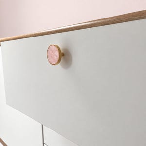 Op de afbeelding: Een wit ladefront met een ronde knop voorzien van een roze, gemarmerde stenen inleg in een goudkleurig metalen frame. De lade bevindt zich onder een horizontale houten rand, wat zorgt voor een strak, vlak oppervlak met één enkel hardware-accent.