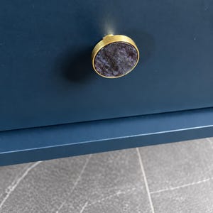 Op de afbeelding: Een ronde meubelknop met een rand van geborsteld goudkleurig metaal en een donkerpaarse, gemarmerde stenen inleg met subtiele sprankelende spikkels, gemonteerd op een gladde, mat marineblauwe ladefront.