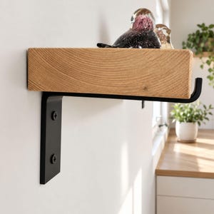 Könnte beinhalten: Ein dickes, helles Holzregal, montiert auf einer mattschwarzen Metallhalterung mit J-förmigem Ende. Das Regal zeigt eine sichtbare Holzmaserung und ist mit zwei schwarzen Schrauben an der Wand befestigt. Eine dunkle Glasvogel-Figur steht darauf.