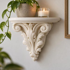 Op de afbeelding: Een wit wandrek met een sierlijke, uitgesneden console-steun met gedetailleerde acanthusbladmotieven. Het platte bovenoppervlak bevat een grijze gespikkelde plantenpot met een hangplant en een glazen kaarshouder met een brandende kaars.