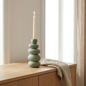 Puede incluir: Un portavelas de color verde salvia con un diseño apilado de formas redondeadas similares a guijarros. El soporte tiene un acabado mate suave y sostiene una vela cónica delgada de color crema con una pequeña llama encendida en la mecha.
