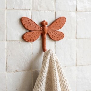 Pode incluir: Um gancho de parede em forma de libélula na cor terracota com padrões detalhados nas asas. O gancho está montado numa parede de azulejos brancos e segura um tecido bege com textura de waffle pendurado na parte inferior do corpo.