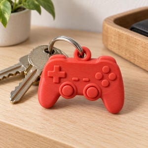 May include: A bright red keychain shaped like a video game controller with a directional pad, two analog sticks, and four action buttons. The controller is attached to a silver metal split ring that holds two standard silver metal keys.