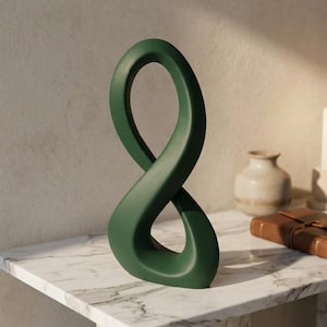 Op de afbeelding: Een donkergroen keramieken sculptuur in de vorm van een doorlopende oneindigheidslus. Het object heeft een gladde, matte afwerking en een hol midden, staat rechtop en heeft een vloeiend, gebogen ontwerp dat een gebalanceerd silhouet vormt.