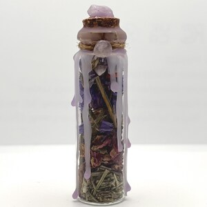 Puede incluir: Un vial de vidrio transparente lleno de flores secas, hierbas y piedras pequeñas, sellado con un tapón de corcho. Cera de color violeta claro gotea sobre la parte superior y los lados del vidrio, con una pequeña piedra violeta en la cima.