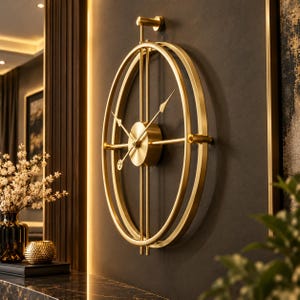 May include: A gold-toned wall clock featuring a double-ring oval frame and a central circular housing. The clock has thin, straight hands and a vertical rod structure, mounted to the wall with a small cylindrical bracket at the top centre.