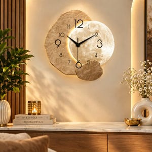 May include: A wall clock featuring a textured, beige, stone-like irregular frame and a circular, illuminated moon-themed face. The clock has black hands and black Arabic numerals from 1 to 12, with a warm light glowing from behind the moon disc.