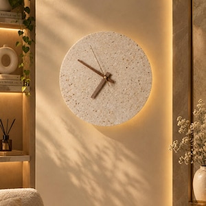 Puede incluir: Un reloj de pared circular con una esfera de color beige claro moteado estilo terrazo y manecillas de madera marrón oscuro. El reloj cuenta con un segundero fino de tono dorado y está montado en una pared con un efecto de luz cálida trasera.