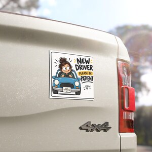 Op de afbeelding: Een vierkante autosticker met een cartoonillustratie van een persoon met een bruine knot die in een blauwe auto rijdt. De tekst luidt NEW DRIVER PLEASE BE PATIENT en op de nummerplaat staat JUST LEARNING, vergezeld van een klein hartsymbool.