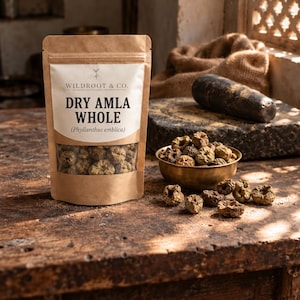 Może przedstawiać: Brązowa papierowa torebka z etykietą Wildroot & Co. Dry Amla Whole (Phyllanthus emblica) zawierająca suszone jagody amla. Obok torebki znajduje się mała mosiężna miseczka wypełniona suszonymi jagodami amla oraz kilka luźnych jagód obok.
