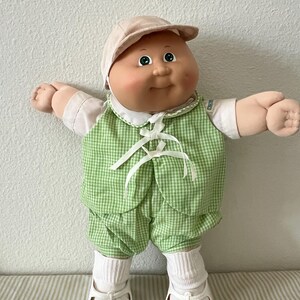 May include: A doll with a round face, green eyes, and dimpled cheeks, wearing a beige cap, a white short-sleeved shirt, and a green and white gingham vest and shorts set. The doll also wears white socks and white shoes with a floral pattern.