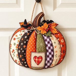 May include: A fabric pumpkin wall hanging featuring patchwork segments in floral, orange, green spiderweb, and purple gingham patterns. It is decorated with a small heart patch, a black witch hat, a bat, and artificial autumn leaves.