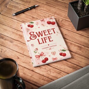 Könnte beinhalten: Ein rechteckiges Notizbuch mit einem hellrosa Einband, verziert mit roten Kirschillustrationen, kleinen Schleifen und Herzen. Der Text SWEET LIFE und CHERRY EDITION ist in Rot auf der Vorderseite gedruckt. Das Notizbuch liegt auf einer Holzoberfläche.