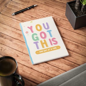 Könnte beinhalten: Ein rechteckiges Notizbuch mit weißem Einband und dem farbigen Text YOU GOT THIS sowie ONE DAY AT A TIME. Das Design enthält kleine Blumen- und Sternillustrationen in Pastellfarben sowie eine hellblaue Wellenlinie am linken Rand.