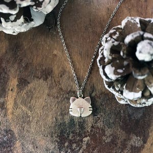 May include: A silver necklace with a small cat charm pendant. The necklace is on a wooden surface with pine cones.