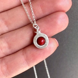 Może przedstawiać: Srebrny łańcuszek z małym, czerwonym wisiorem w kształcie granatu. Wisiorek jest wykonany ze srebra i ma fakturowaną powierzchnię.