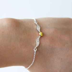 Puede incluir: Una pulsera de plata con un dije de oro en forma de un par de alas. Las alas están detalladas con líneas y el dije es una cuenta redonda.