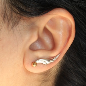 May include: A silver ear cuff with a gold ball at the end. The cuff is shaped like a wing and is worn in the upper part of the ear.