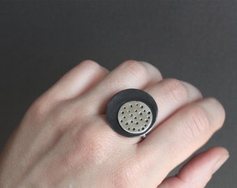 May include: A large, round ring with a two-tone design. The outer circle is dark grey, and the inner circle is silver with small, evenly spaced holes. The ring is worn on a finger.
