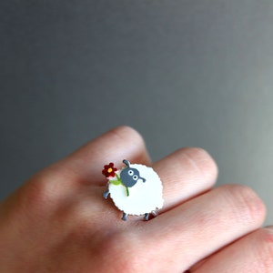 May include: A white sheep ring with a black face and a red flower. The sheep is holding the flower in its mouth and has a green stem.