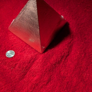 Puede incluir: Una pirámide de color cobre metálico con un acabado de superficie texturizado. La pirámide está colocada junto a una pequeña moneda circular de color plateado para dar escala, lo que proporciona una referencia visual clara del tamaño.