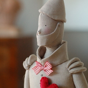 Puede incluir: Una muñeca de tela hecha a mano del Hombre de Hojalata del Mago de Oz. La muñeca lleva un sombrero de tela beige, una camisa de tela beige con una corbata de lazo roja y blanca, y un corazón de fieltro rojo. La muñeca tiene un collar de cadena y el texto "Nastri & Rocchetti Country" está impreso en la camisa de la muñeca.