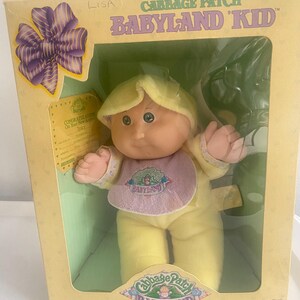 Op de afbeelding: Een Cabbage Patch Babyland Kid pop in een gele outfit met een paars slabbetje, verpakt in een gele doos met een paarse strik. De doos bevat de tekst Cabbage Patch Babyland Kid, een afbeelding van een geboortecertificaat en het Coleco-logo.