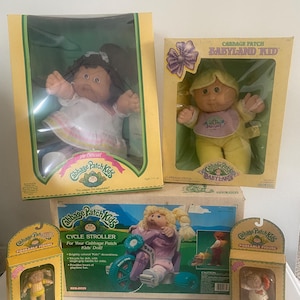 Könnte beinhalten: Eine Sammlung von Cabbage Patch Kids Spielzeug, bestehend aus zwei großen Puppen in gelben Kartons, einem Cycle Stroller Karton mit einer Puppe auf einem Dreirad und zwei kleinen Schachteln mit beweglichen Figuren, inklusive Originalverpackung.
