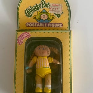 May include: A Cabbage Patch Kids poseable figure in its original yellow packaging. The doll has brown yarn hair in braids, a yellow and white checkered top, and yellow shorts. The box features the text Cabbage Patch Kids, Second Edition, and Hello, My Name Is Sue Georgia.