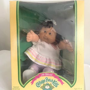 May include: A Cabbage Patch Kids doll with dark brown hair and a white dress featuring pink and yellow stripes, packaged in a yellow box with green trim. The box displays the text The Official Cabbage Patch Kids, I'm unique. I'm adoptable!, and Ages 3 & up.