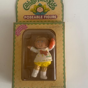 May include: A Cabbage Patch Kids poseable figure in its original packaging. The doll has blonde hair, blue eyes, and wears a white dress with a red bow and yellow bloomers. The box features the text Cabbage Patch Kids, Poseable Figure, and Second Edition.