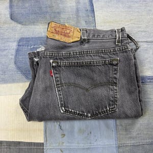 May include: A pair of charcoal grey denim jeans featuring a back pocket with a red tab, belt loops, and a tan leather patch on the waistband printed with Levi Strauss & Co. 501 W 40 L 30. The denim fabric shows signs of wear with light fading.