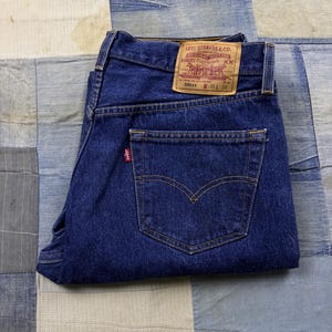 May include: Dark blue denim jeans featuring a tan leather patch on the back waistband with the text Levi Strauss & Co. Original Riveted Quality Clothing XX 501xx W 36 L 36. A red tab is attached to the back pocket with the Levi's brand name.