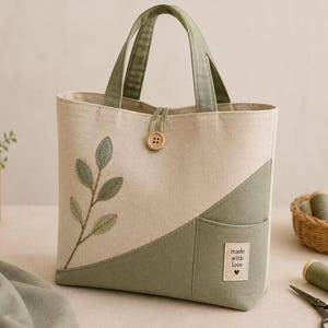 Puede incluir: Un bolso de tela en color beige y verde salvia con dos asas, cierre de botón de madera y un diseño bordado de rama con hojas. El frente incluye un bolsillo diagonal con una etiqueta rectangular que dice made with love y un corazón.