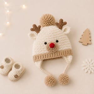 Puede incluir: Un gorro de ganchillo color crema con diseño de reno, astas marrones, nariz roja y tres pompones beige. El gorro tiene orejeras y se acompaña de un par de patucos de ganchillo a juego en color crema con pequeños botones marrones.