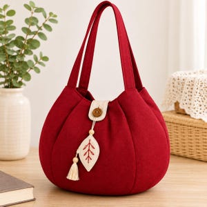 Puede incluir: Un bolso de tela rojo intenso con silueta redondeada y plisada, con dos asas. El bolso cuenta con un cierre de lengüeta beige asegurado por un botón marrón, adornado con un colgante en forma de hoja y una pequeña borla decorativa.