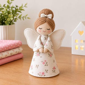 Puede incluir: Una muñeca ángel de tela con cabello de lana marrón claro recogido en un moño, adornado con un pequeño lazo blanco. La muñeca viste un vestido color crema con estampado floral rosa, alas de tela blanca y rasgos faciales bordados.