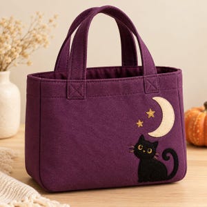 Puede incluir: Un bolso tote de lona en color morado oscuro con dos asas resistentes y un diseño de aplique bordado en la parte frontal. El diseño incluye un gato negro sentado bajo una luna creciente color crema y dos pequeñas estrellas doradas.