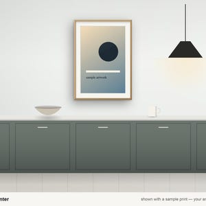 May include: A framed rectangular print featuring a dark circle and a horizontal white line over a gradient background, hanging above a dark green kitchen counter with a bowl, a mug, and a black pendant light fixture.