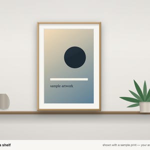 May include: A rectangular wooden picture frame containing a print with a gradient background, a dark blue circle, a horizontal white line, and the text "sample artwork" in black, positioned on a shelf next to a small vase and a plant.