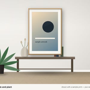May include: A rectangular wooden frame containing a print with a gradient background, a dark circle, a horizontal white line, and the text "sample artwork". The frame sits on a dark brown console table next to a vase and a green plant.