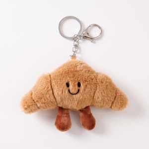 Può includere: Un portachiavi a forma di croissant in peluche con una consistenza morbida e soffice color marrone chiaro, un semplice viso sorridente ricamato in nero e due piccole gambe marroni pendenti. È fissato a un anello in metallo argentato e moschettone.