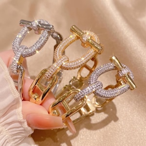Puede incluir: Tres pulseras de eslabones con cierre de barra y argolla. Una pulsera es de tono plateado, otra de tono dorado y una combina tonos dorados y plateados. Cada pulsera está incrustada con pequeñas piedras transparentes y brillantes.